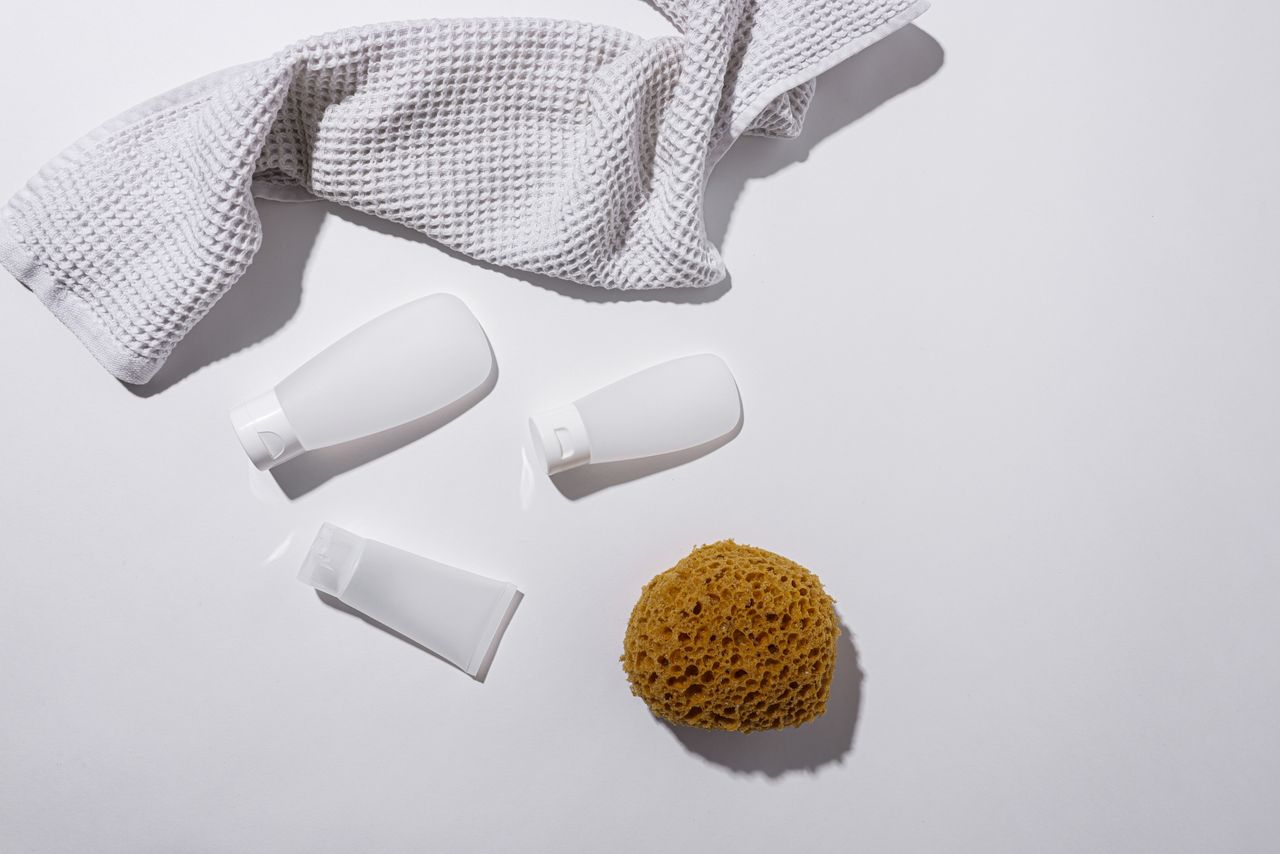 A clean, minimalist arrangement of bathroom essentials including bottles, a towel, and a sponge on a white background.
